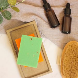 Flat lay of natural skincare products including soap bars, oils, and brush on soft fabric.